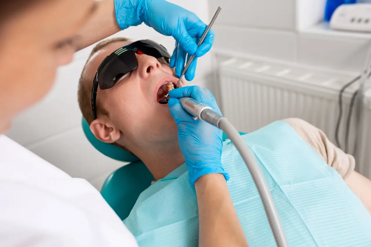 Young man visiting the dental office. Dentist hands in protective gloves using ultrasound to clean patient's teeth. Closeup of open mouth.