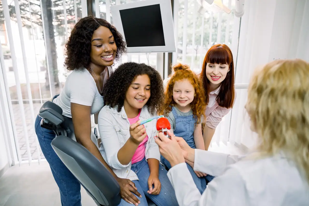 Dental care, teeth hygiene and check up. Back view of female dentist showing teeth model to African and Caucasian teenagers girls, visiting modern dental clinic with their young multiracial mothers