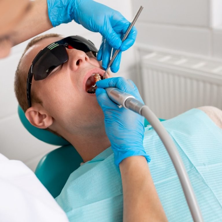 Young man visiting the dental office. Dentist hands in protective gloves using ultrasound to clean patient's teeth. Closeup of open mouth