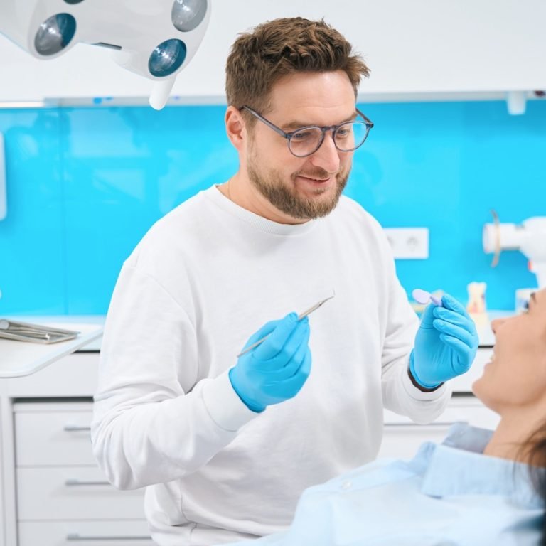 Friendly doctor stomatologist holding mouth mirror and sickle probe to to find tooth decay or other potential oral problems in female patient, check-up