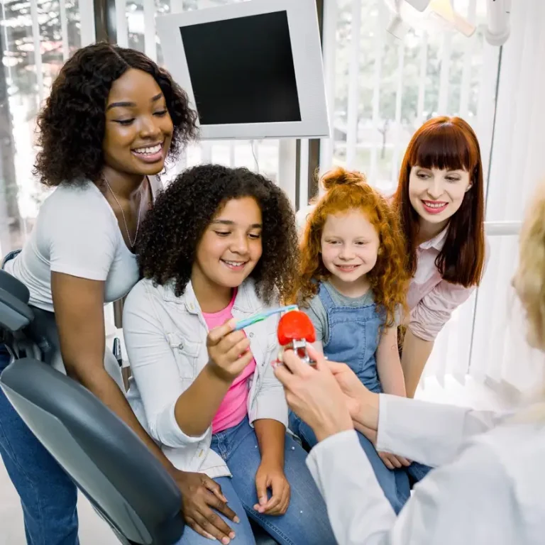 Dental care, teeth hygiene and check up. Back view of female dentist showing teeth model to African and Caucasian teenagers girls, visiting modern dental clinic with their young multiracial mothers